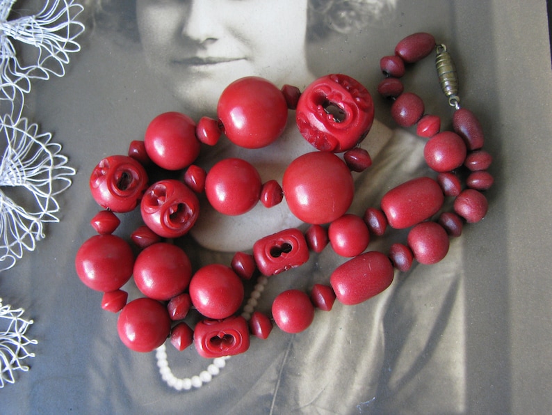 Peut inclure: Un collier de perles rouges avec des perles en bois sculpt&eacute;es. Le collier est compos&eacute; de deux rangs de perles, un rang est plus grand que l'autre. Les perles sont d'une couleur rouge fonc&eacute; et ont une finition brillante.