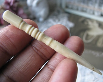 May include: A small, white, bone-like tool with a pointed tip and a decorative, ridged section. The tool is held in a hand.