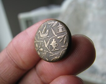 May include: A round, brass button with an intricate design featuring a bird, a teapot, and other floral motifs.