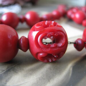 Peut inclure: Gros plan d'un collier de perles rouges avec des d&eacute;tails sculpt&eacute;s complexes sur l'une des perles. Les perles sont reli&eacute;es par une fine cha&icirc;ne m&eacute;tallique.