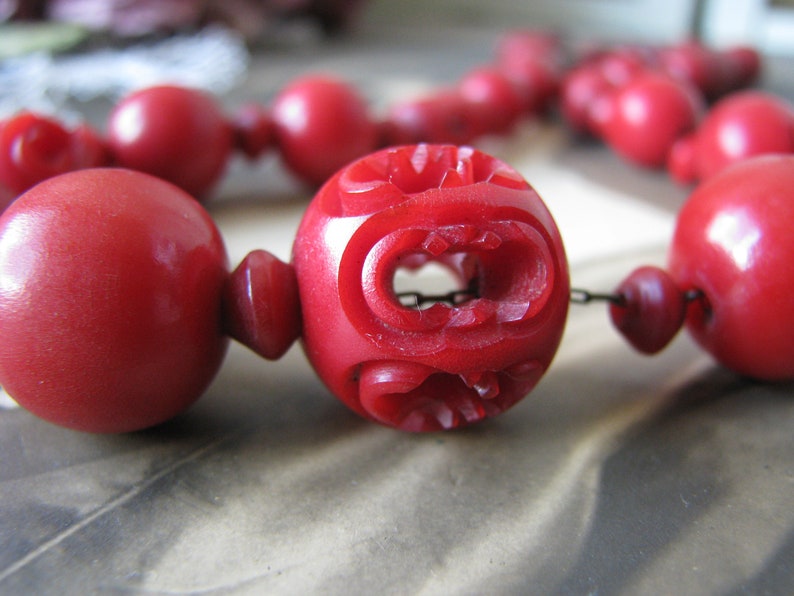 Peut inclure: Gros plan d'un collier de perles rouges avec des d&eacute;tails sculpt&eacute;s complexes sur l'une des perles. Les perles sont reli&eacute;es par une fine cha&icirc;ne m&eacute;tallique.