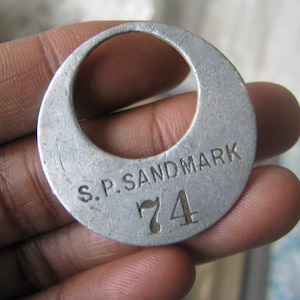 May include: A silver metal tag with the text "S.P. SANDMARK" and the number "74" engraved on it.
