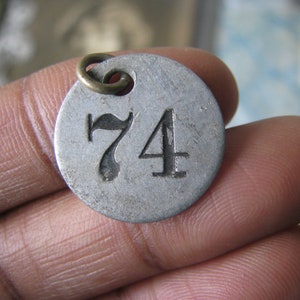 May include: A round, silver-colored metal tag with the number 74 stamped on it. The tag has a small hole at the top with a brass-colored ring attached. The tag is held in a person's hand.