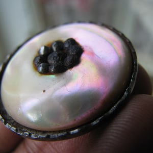 Puede incluir: Un anillo plateado con una gran piedra blanca nacarada iridiscente engastada en el centro. La piedra tiene un grupo de pequeños acentos redondos negros en el centro.