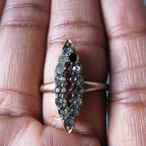 Puede incluir: Un anillo de oro con un engaste ovalado puntiagudo. El engaste est&aacute; lleno de peque&ntilde;as piedras preciosas transparentes y rojas.