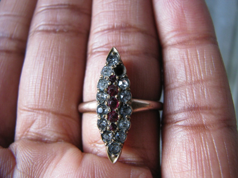 Puede incluir: Un anillo de oro con un engaste ovalado puntiagudo. El engaste est&aacute; lleno de peque&ntilde;as piedras preciosas transparentes y rojas.
