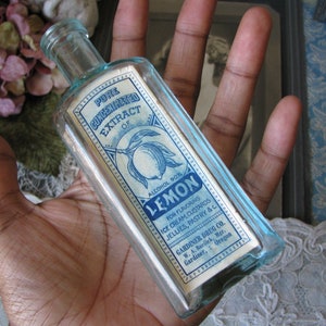 May include: A clear glass bottle with a blue label that reads "Pure Concentrated Extract of Lemon, Alcohol 90%". The label also includes instructions for using the extract for flavoring ice cream, custards, jellies, pastry, and other foods. The bottle is held in a hand.