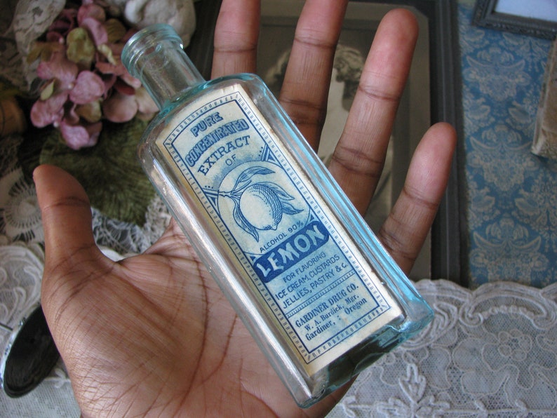 May include: A clear glass bottle with a blue label that reads "Pure Concentrated Extract of Lemon, Alcohol 90%". The label also includes instructions for using the extract for flavoring ice cream, custards, jellies, pastry, and other foods. The bottle is held in a hand.