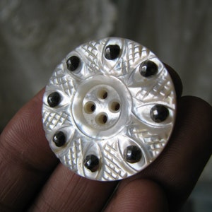 Puede incluir: Un botón blanco de nácar con un centro de cuatro agujeros y un diseño decorativo de ocho acentos negros alrededor del borde.