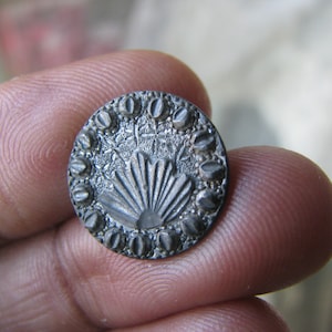 May include: A silver-toned button with a decorative design featuring a fan-shaped motif surrounded by a border of small, raised dots.