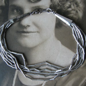 Op de afbeelding: Een zilveren ketting met meerdere strengen kleine, cilindrische kralen. De ketting heeft een eenvoudige sluiting.