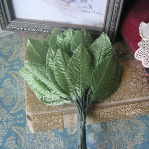 May include: A bunch of green fabric leaves, tied together at the base, with a brown book in the background.