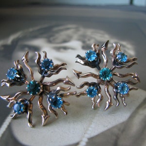 May include: A pair of vintage earrings with a gold tone metal base and blue rhinestones. The earrings are shaped like a starburst with a swirling design.