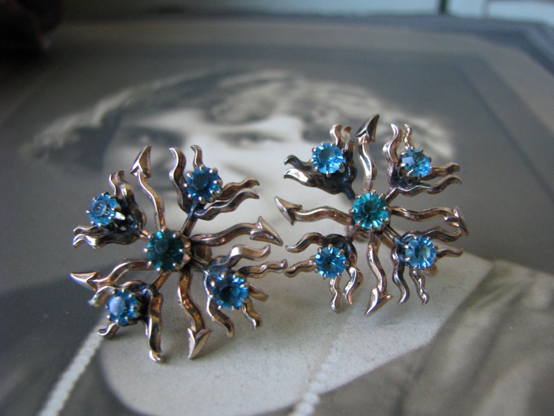 May include: A pair of vintage earrings with a gold tone metal base and blue rhinestones. The earrings are shaped like a starburst with a swirling design.