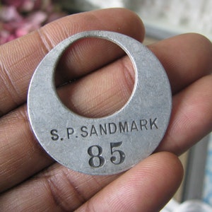 May include: A silver metal tag with the text "S.P. SANDMARK 85" engraved on it.