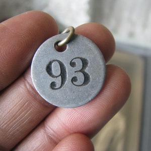 May include: A silver metal tag with the number 93 engraved on it. The tag has a hole at the top and is attached to a brass ring.