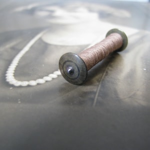 May include: A small spool of brown thread with a metal casing. The spool is lying on a vintage photograph of a woman wearing a pearl necklace.