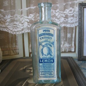 May include: A vintage glass bottle with a blue and white label that reads "Pure Concentrated Extract of Lemon, Alcohol 90%". The label also includes instructions for using the extract for flavoring ice cream, custards, jellies, pastry, and other foods. The bottle is sitting on a table with a lace tablecloth and a framed picture.