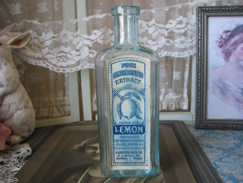 May include: A vintage glass bottle with a blue and white label that reads "Pure Concentrated Extract of Lemon, Alcohol 90%". The label also includes instructions for using the extract for flavoring ice cream, custards, jellies, pastry, and other foods. The bottle is sitting on a table with a lace tablecloth and a framed picture.