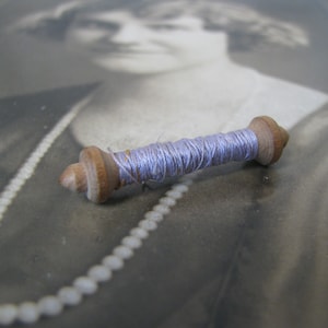 May include: A wooden spool of light blue thread, lying on a black and white photograph of a woman wearing a pearl necklace.