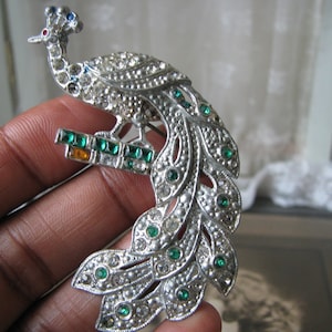 Może przedstawiać: Srebrna broszka w kształcie pawia z zielonymi i przezroczystymi cyrkoniami. Paw jest osadzony na gałęzi z zielonymi i pomarańczowymi cyrkoniami.