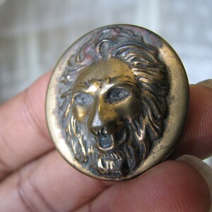 Pode incluir: Um botão de metal dourado com um design detalhado de um rosto de leão. A juba do leão é esculpida de forma intrincada e o botão tem um acabamento ligeiramente desgastado.