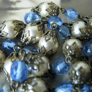 May include: A close-up of a necklace with alternating white pearl and blue glass beads. The pearl beads are surrounded by ornate metalwork.