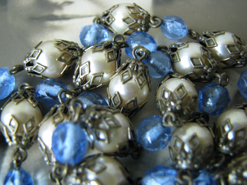May include: A close-up of a necklace with alternating white pearl and blue glass beads. The pearl beads are surrounded by ornate metalwork.