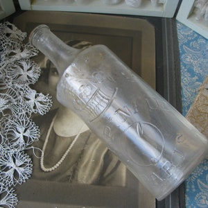 May include: A clear glass bottle with a textured surface and embossed lettering. The bottle is empty and has a cork stopper. The bottle is sitting on a white lace doily.