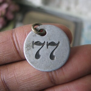 May include: A silver metal tag with a black number 77 on it. The tag has a small brass loop at the top.