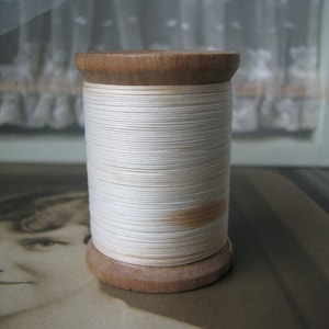 Op de afbeelding: Een spoel met witte draad gewikkeld om een houten spoel. De draad is strak gewikkeld en heeft een licht gebroken witte kleur. De spoel is gemaakt van natuurlijk hout en heeft een gladde afwerking.