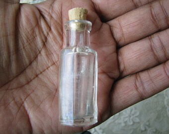Puede incluir: Una pequeña botella de vidrio transparente con un tapón de corcho. La botella está vacía y tiene una superficie ligeramente texturizada.