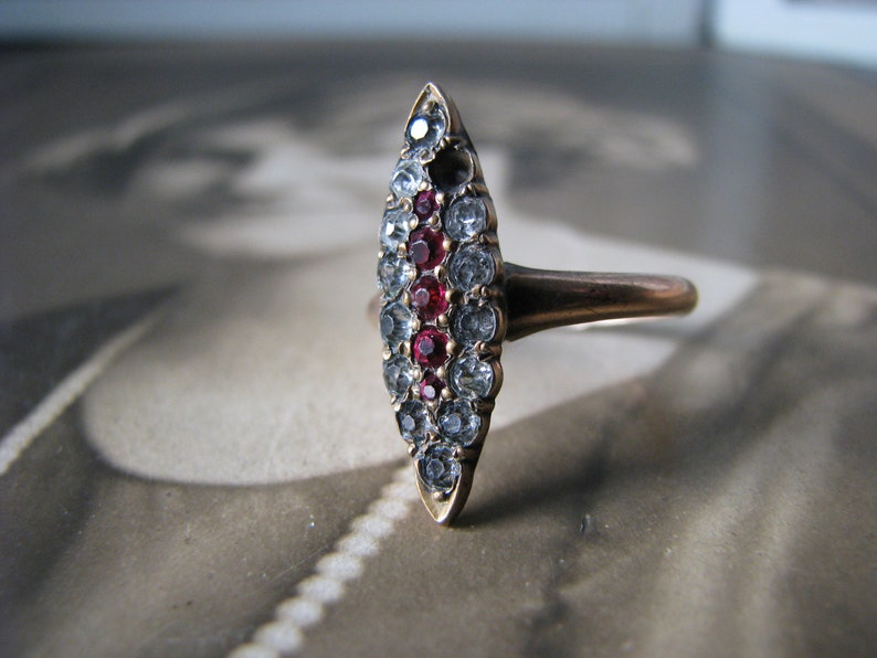 Puede incluir: Un anillo de oro con un engaste ovalado puntiagudo. El engaste est&aacute; lleno de peque&ntilde;as piedras transparentes y una fila de piedras rojas en el centro.
