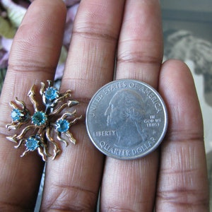 May include: A pair of gold tone earrings with blue rhinestones set in a sunburst design. The earrings are shown next to a US quarter for size comparison.