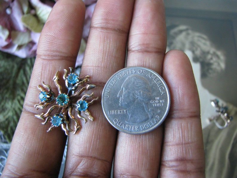 May include: A pair of gold tone earrings with blue rhinestones set in a sunburst design. The earrings are shown next to a US quarter for size comparison.