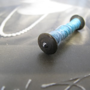 May include: A small spool of light blue thread with a metal cap. The spool is lying on a gray surface.