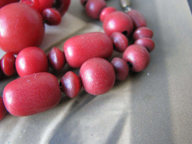 Peut inclure: Gros plan d'un collier de perles en bois rouge. Les perles sont de diff&eacute;rentes tailles et formes, et le collier est enfil&eacute; sur un cordon fonc&eacute;.
