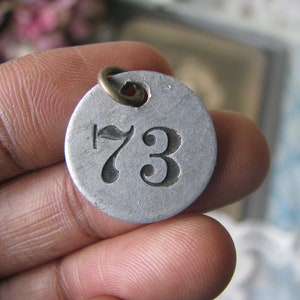 May include: A silver metal tag with the number 73 engraved on it. The tag has a small brass loop at the top.