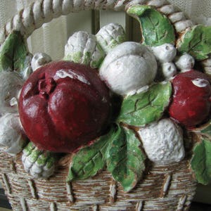 May include: A white ceramic basket filled with red and white fruit, green leaves, and white flowers. The basket is decorated with a rope-like design around the rim.