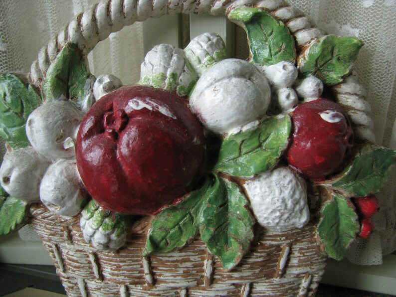 May include: A white ceramic basket filled with red and white fruit, green leaves, and white flowers. The basket is decorated with a rope-like design around the rim.