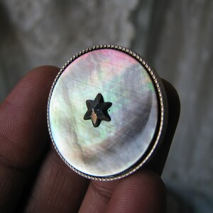 May include: A round, silver-rimmed button with a mother-of-pearl face. The button has a black star-shaped design in the centre.