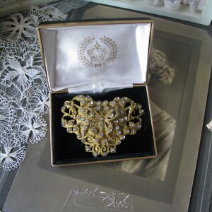 May include: A gold-toned brooch in the shape of a heart with a floral design and clear rhinestones. The brooch is in a black velvet-lined box with a white satin interior. The box has a gold-toned crown logo on the lid.