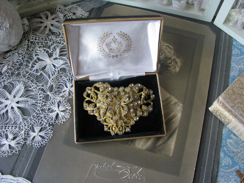 May include: A gold-toned brooch in the shape of a heart with a floral design and clear rhinestones. The brooch is in a black velvet-lined box with a white satin interior. The box has a gold-toned crown logo on the lid.
