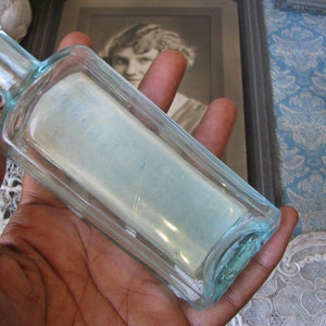 May include: A clear glass bottle with a square shape and a light blue tint. The bottle is empty and has a smooth, polished surface.