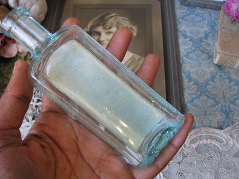 May include: A clear glass bottle with a square shape and a light blue tint. The bottle is empty and has a smooth, polished surface.