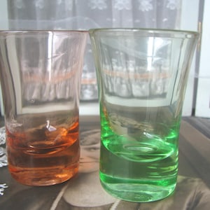 Può includere: Vengono mostrati due bicchierini vintage, uno arancione e uno verde. I bicchieri sono trasparenti con una leggera sfumatura di colore sul fondo. I bicchierini sono su un tavolo davanti a una finestra con tende di pizzo bianco.