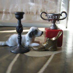 Puede incluir: Una figurita de porcelana blanca y negra de un perro acostado junto a un candelero plateado, una copa de trofeo plateada sobre un cubo rojo y un zapato dorado.