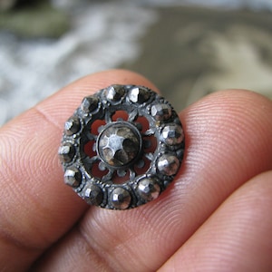 Puede incluir: Un botón de plata con un diseño decorativo y un centro elevado. El botón está rodeado por un anillo de pequeños puntos en relieve.