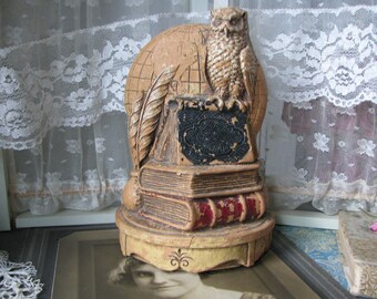 Puede incluir: Un pisapapeles vintage de madera pintada con forma de búho. El búho está posado sobre una pila de libros con un globo terráqueo detrás. El pisapapeles está pintado en tonos marrones y dorados.