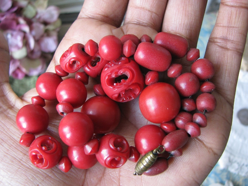 Peut inclure: Un collier de perles rouges avec des perles sculpt&eacute;es et un fermoir en m&eacute;tal. Le collier est compos&eacute; de perles rouges de diff&eacute;rentes tailles et formes.